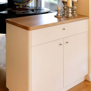 Painted beige satin-finished cabinet with rounded edges, a wide drawer with 2 doors below, with a silver knob handle and thick lacquered wooden top with 5 chunky silver candle holders, and a piano with a globe in the background