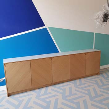 A wooden sideboard with original grooved corners on the fronts, forming handles, background wall, and base with blue and turquoise shapes
