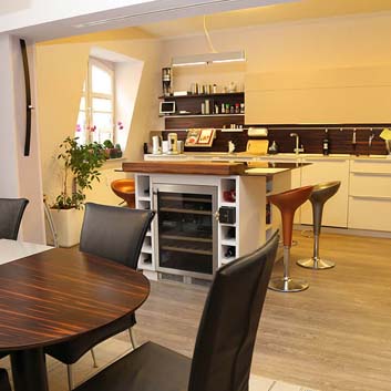 Eine große Küche mit Wand-zu-Wand-weißen, eleganten Schränken, schwarzem Backsplash und einer Mittelblockinsel mit eingebautem Weinkühlschrank und Barhockern