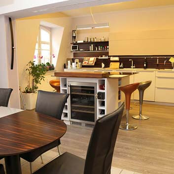 A large kitchen with wall-to-wall white elegant cabinets, black backsplash, and a central island with a built-in wine refrigerator and bar stools