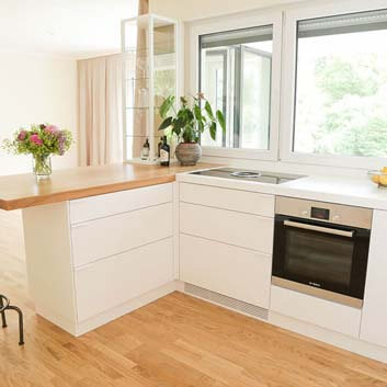 L-shaped white kitchen with shorter side featuring a chunky natural wood top serving as a breakfast table, with a bar stool, Bora stove with integrated extractor, and Bosch oven, and a showcase with glass shelves on the countertop