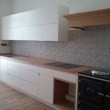 Wall-to-wall kitchen with painted light fronts, wooden countertop with Bora induction stove, an open wooden shelf for wood next to a traditional wood stove