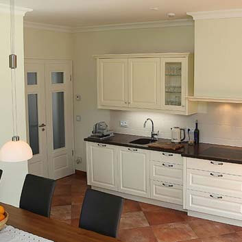 Kitchen with classically grooved white kitchen fronts with handlebars, dark countertop, and white wall cabinets with full doors and glass doors, hiding/storage space behind a sliding door