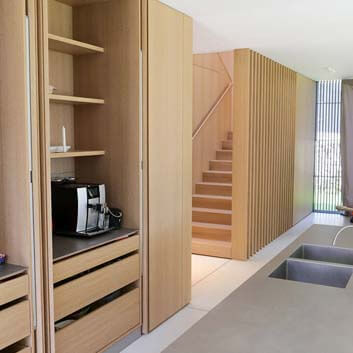 Kitchen with revolving doors hiding a functional kitchen with oak drawers, shelves, a coffee machine, partially visible modern solid wood staircase, and partially visible kitchen island with Bora stove and Silvertouch countertop.