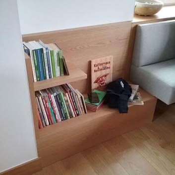 Built-in bench with gray padded seat and backrest, featuring an original bookshelf solution on the side of the bench.