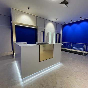 New empty cosmetic salon, with white Corian counter, featuring golden ABET LAMINATI element, walnut wood slats on both sides, and LED lighting under the counter, behind a golden frame with light and blue background, Occhio Sento and Mito ceiling lights.