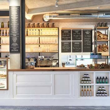 Restaurant in Hamburg, white counter with chunky wooden top, featuring glass showcases displaying food, tall open shelves showcasing wine bottles and freshly baked bread.